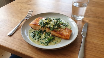 Un plato de salmón con salsa cremosa de espinaca, ajo y limón sobre una mesa de madera clara, acompañado de un tenedor, un cuchillo y un vaso de agua.