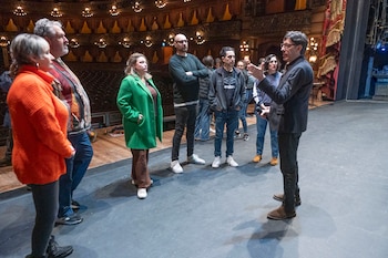 Un grupo de personas se reúne en el escenario de un teatro ornamentado; un hombre de traje negro habla y gesticula frente a ellos
