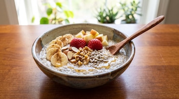 Bol de avena con chía, rodajas de banano, fresas, cubos de manzana, almendras, nueces, semillas de girasol, miel, canela y una cuchara de madera sobre mesa de madera.