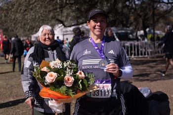Una mujer deportista con medalla y dorsal sostiene un ramo de rosas junto a una mujer mayor apoyada en un bastón al aire libre