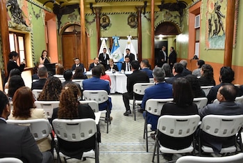 Varias personas sentadas de espaldas observan a cuatro funcionarios en una mesa con banderas de Guatemala al fondo