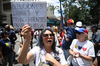 Mujer con gafas de sol y camiseta blanca sostiene un cartel que dice 'ELECCIONES LIBRES Y TRANSPARENTES EN VENEZUELA' en medio de una multitud