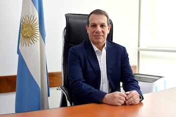Un hombre con saco azul y camisa blanca permanece sentado frente a un escritorio junto a la bandera de Argentina