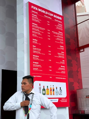Un hombre en camisa blanca y corbata negra frente a un cartel rojo con el menú de bebidas de la Copa Mundial 2026, mostrando precios de licores y agua