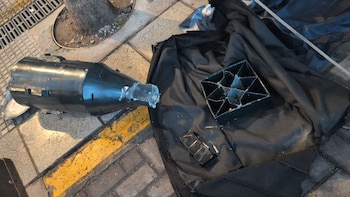 Objeto metálico oscuro, cilíndrico, parecido a una bomba, yace en la acera junto a un desagüe y un árbol, con otros fragmentos metálicos sobre una tela negra