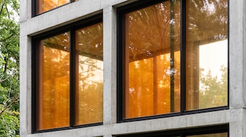Dos ventanales grandes de color ámbar-naranja con marcos negros en una fachada de hormigón. Se observan reflejos tenues de árboles verdes.