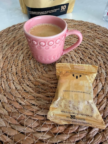 Taza de cerámica rosa con café, un alfajor Havanna en envoltorio dorado, un individual trenzado y un paquete de diez alfajores