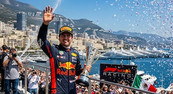Sergio "Checo" Pérez sonriente, ondeando la mano y sosteniendo un trofeo, con confeti cayendo. De fondo, el puerto de Mónaco, yates y edificios.