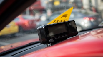 Taxímetro negro apagado en el tablero de un taxi rojo, con un logo triangular amarillo de taxi en el parabrisas y fondo desenfocado de coches urbanos.