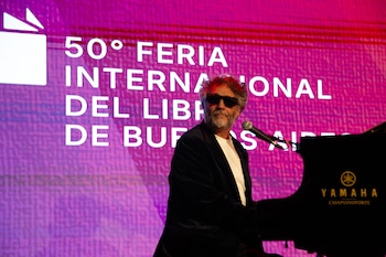 Fito Páez, con gafas de sol y barba, sentado frente a un piano negro Yamaha, con una pantalla LED púrpura de fondo que muestra "50° FERIA INTERNACIONAL DEL LIBRO"