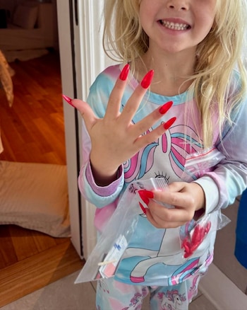 Una niña con prenda de unicornio enseña sus manos con uñas postizas rojas muy largas y sostiene pequeñas bolsas transparentes con accesorios