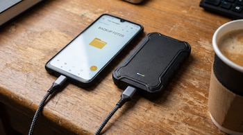 Un teléfono móvil conectado por cable a un disco de almacenamiento externo sobre una mesa de madera junto a un vaso de café.