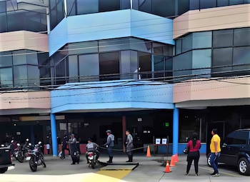 Fachada de un edificio comercial de cristal azul y beige con personas, motocicletas, conos de tránsito y un automóvil en la entrada