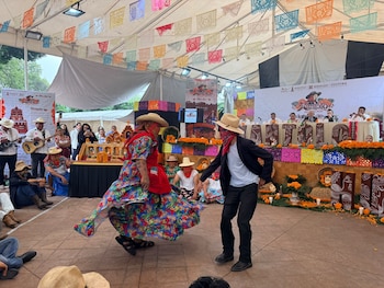 Dos personas con máscaras, sombreros y trajes folclóricos bailan bajo una carpa decorada con papel picado, flores de cempasúchil y estandartes