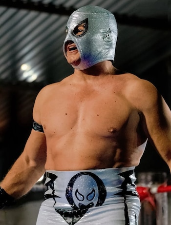 Hombre musculoso enmascarado con máscara plateada, torso desnudo y pantalón de lucha blanco y negro, en un ring