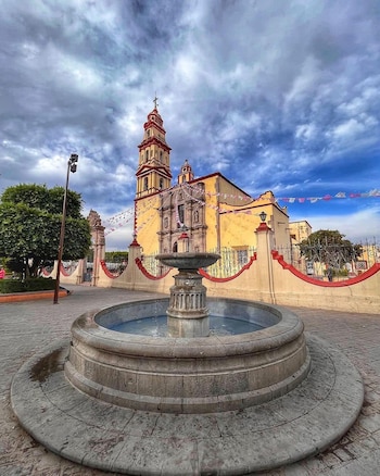 Fuente circular de piedra en el primer plano de una plaza con una iglesia amarilla y un cielo nublado
