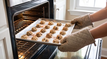 Manos enguantadas sujetan una bandeja de galletas de avena, chocolate y canela crudas sobre un horno encendido con la puerta abierta.