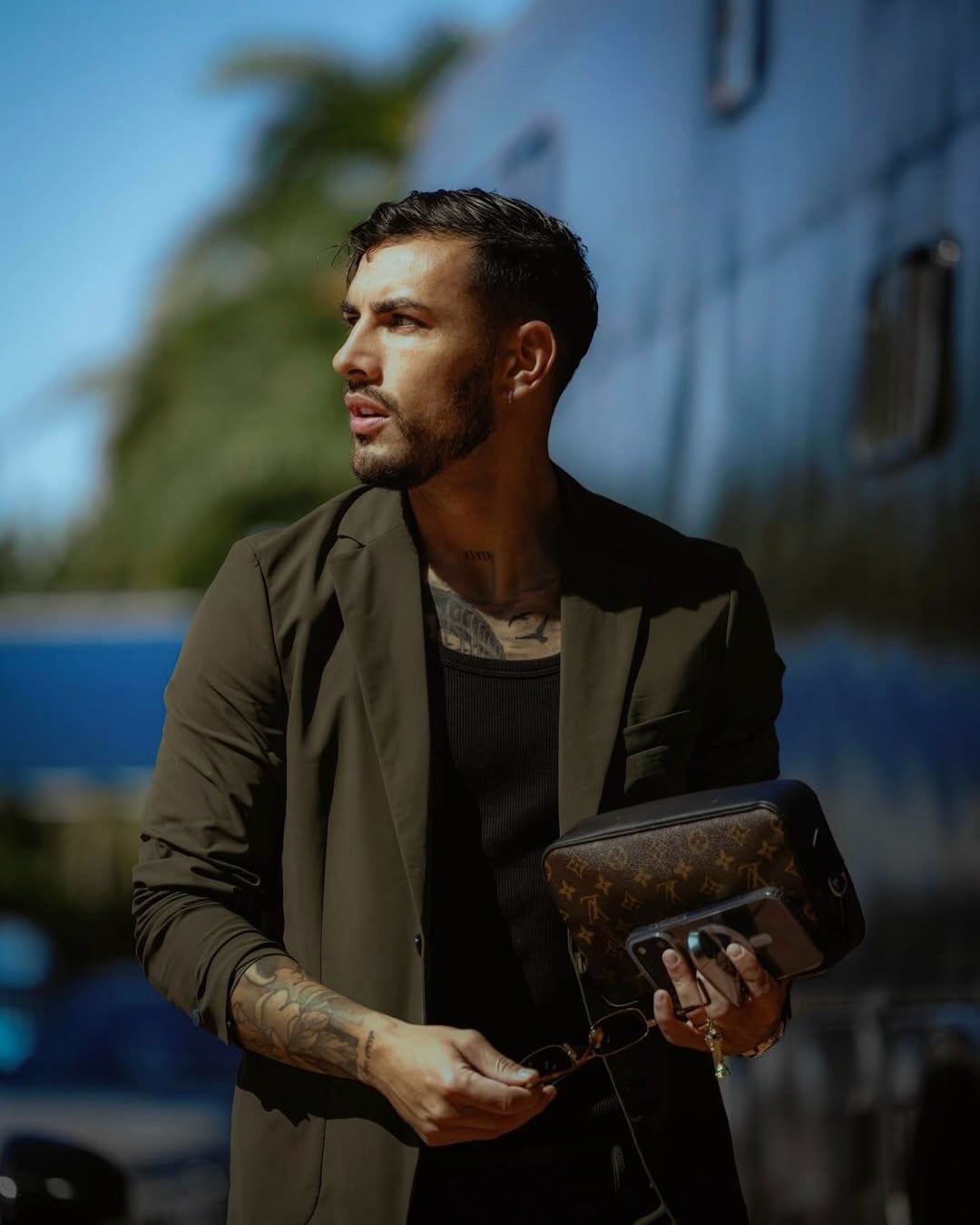 Leandro Paredes exhibe un look moderno y urbano, combinando un blazer verde oscuro, camiseta negra, y accesorios de lujo como un bolso Louis Vuitton, gafas de sol y un teléfono (Instagram Selección)