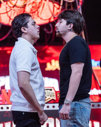 Dois jovens se encarando em uma cena. Um está vestindo uma camisa pólo branca e o outro uma camiseta preta com corrente dourada e relógio.