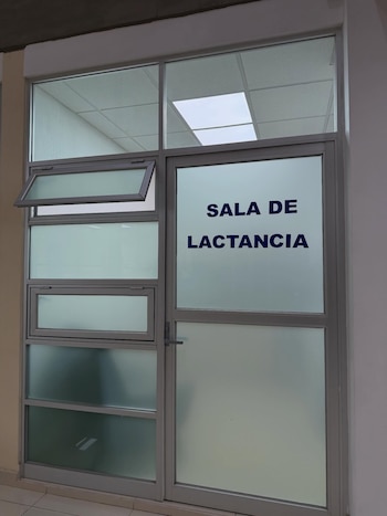 Vista exterior de una sala de lactancia. La puerta de vidrio esmerilado tiene el texto 'SALA DE LACTANCIA' en azul. Ventanas superiores abiertas