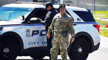 Un militar de la Fuerza Aérea con uniforme de camuflaje y boina negra está de pie frente a un coche patrulla blanco y azul. Un oficial de policía está al fondo