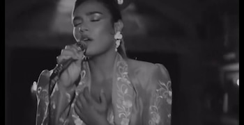 Plano medio en blanco y negro de una mujer que canta ante un micrófono con una mano en el pecho y los ojos cerrados