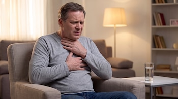 Un hombre de mediana edad con camisa gris se sujeta el cuello y el pecho con ambas manos, sentado en un sillón junto a una mesa con un vaso de agua.