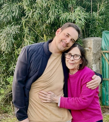 Un hombre sonriente abraza a una mujer con gafas y suéter rosa en un espacio exterior con vegetación