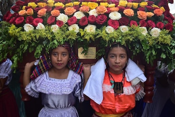 Dos niñas jóvenes con trajes tradicionales cargan una gran plataforma decorada con muchas rosas rojas, amarillas, naranjas y blancas, y follaje verde