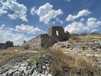 Ruinas de una estructura de piedra, terreno árido con rocas y hierba seca, cielo azul con nubes blancas y dispersas