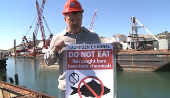 Un hombre con casco rojo sostiene un cartel que advierte en inglés sobre peces contaminados con químicos tóxicos en un puerto con grúas al fondo