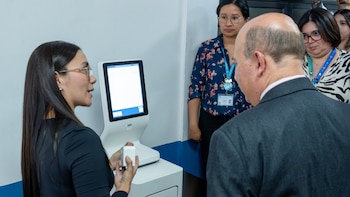 Una mujer con gafas y cabello oscuro sostiene un dispositivo blanco frente a un kiosco digital, hablando con un hombre calvo de espaldas; otras personas observan