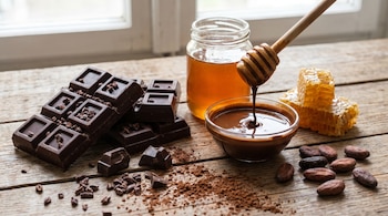 Vista aérea de barras de chocolate oscuro, granos de cacao, miel líquida en un tarro y en un cuenco, panales de abeja y un agitador de miel sobre madera.
