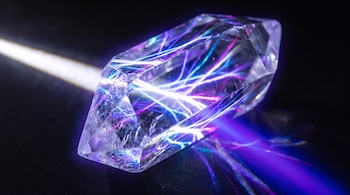 Cristal transparente facetado sobre fondo oscuro. Un haz de luz blanca incide por un lado y proyecta rayos de luz de colores azul y violeta.