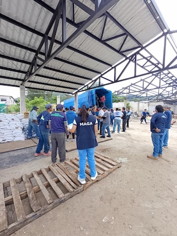 Varias personas bajo una estructura de techo metálico, un camión cubierto de lona azul, sacos apilados y palés de madera en el suelo