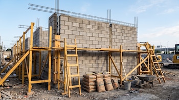 Muro de bloques de concreto gris con vigas de acero, andamios de madera, sacos de cemento y una carretilla en una obra al aire libre.
