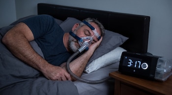 Hombre durmiendo con mascarilla facial y tubo conectados a una máquina CPAP. Despertador digital en mesita de noche muestra la hora 3:17.