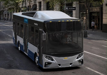 Autobús moderno de colores gris, azul y negro sobre una calle de asfalto, con edificios y un cartel que indica Avenida de Mayo al fondo