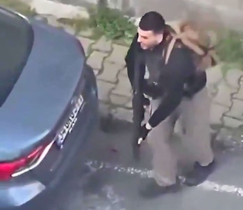 Un hombre con una mochila y ropa oscura agachado junto a la parte trasera de un coche gris oscuro en un suelo adoquinado