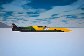 Vehículo de velocidad terrestre JCB Hydromax amarillo y verde con bandera del Reino Unido en la parte trasera, sobre un suelo blanco y plano bajo un cielo azul con nubes