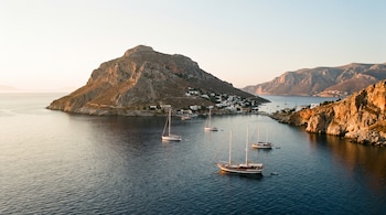 Panorámica de una isla rocosa con casas blancas en su ladera, rodeada por el mar con cinco veleros anclados y montañas distantes bajo un cielo claro.