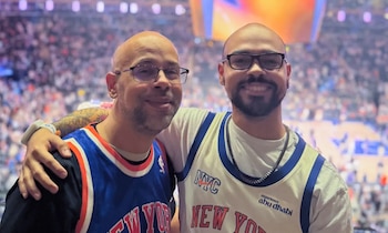 Dos hombres calvos con gafas y camisetas de baloncesto de los Knicks sonríen y se abrazan, con una multitud borrosa de un estadio de fondo