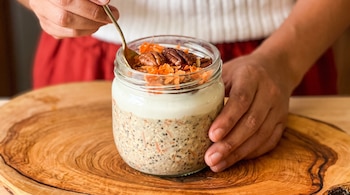 Un frasco de vidrio con avena, chía, yogur, zanahoria rallada y nueces, sostenido por dos manos junto a una cuchara sobre una tabla de madera.