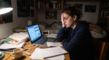 Una joven sentada frente a una computadora portátil abierta, con libros de texto, cuadernos y un reloj digital sobre un escritorio de madera.