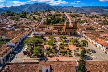 Vista aérea de una plaza con árboles, una iglesia de piedra con dos torres y casas con tejados rojos frente a las montañas.