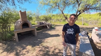 Un hombre con una camiseta oscura que dice WILD THING de pie junto a un horno de barro artesanal y varios árboles.