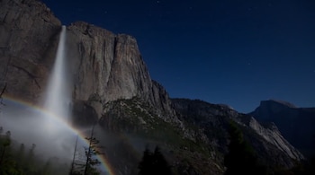Una cascada cae por una imponente pared rocosa bajo un cielo nocturno estrellado, con un tenue arcoíris lunar visible en la base de la cascada