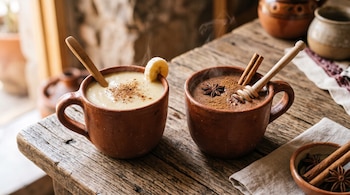 Dos tazas de barro con bebidas calientes de plátano y chocolate decoradas con canela y anís sobre una mesa de madera.
