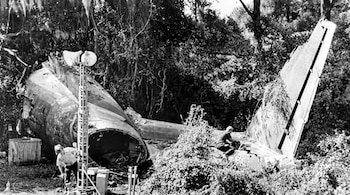 Restos de avión en un área boscosa. Partes del fuselaje y la cola de una aeronave destrozada. Dos hombres y una torre con focos entre árboles y arbustos.