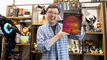 Hombre con lentes y camisa a cuadros azul sostiene libro con ilustración en la portada. Fondo con estantes, figuras de videojuegos, trofeos y un ordenador con luces